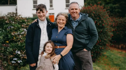 A family photo with a woman, a man a little girl and a teenage boy. They are outside. The mum and daughter are wearing dresses. The dad and teenager have jackets on. Bushes are behind them.