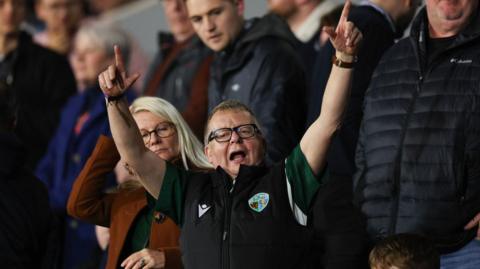A man with brown hair and glasses in a black top in a crowd of people with both of his hands in the air