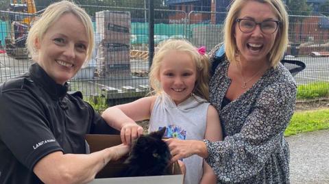 PC Liz Harrison holding a box containing the black cat, with Caroline and her daughter smiling and stroking the cat