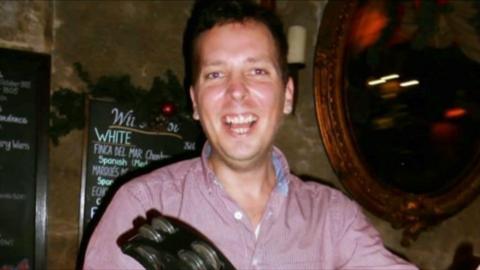 A man is looking at the camera wearing a red shirt. He is smiling. He appears to be a in a dark pub.