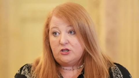 Ms Long speaking as she looks to her left, she has long ginger hair, pink lipstick and is wearing a black and white patterned top with a silver chain necklace.