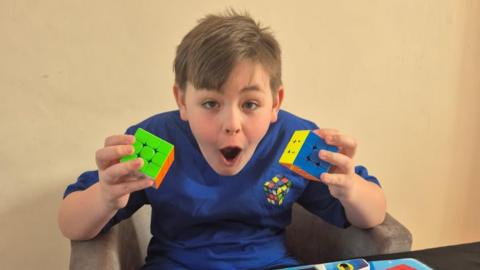 A young boy with short light brown hair swept down over his foreheard holds two Rubik's Cubes and looks enthusiastically at the camera. He is wearing a blue t-shirt with a picture of a Rubik's Cube on it.