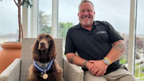 Mark smiles at the camera as he sits on a chair next to Jerry who is wearing his medal