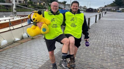Radio presenter James Dundon and business owner Paul Stacey walking along the quayside in Bude in Cornwall. They are both wearing Children in Need branded fluorescent tabards,  and black shorts. They are smiling and Mr Stacey is carry a big, yellow Pudsey bear.