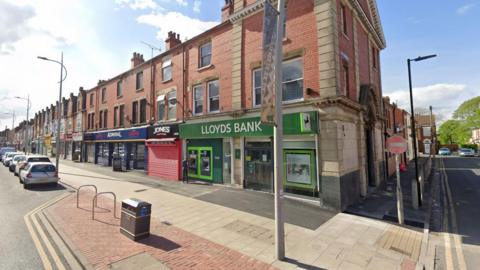 A street view of Hessel Road in Hull. It shows a Lloyds bank, a shop called Jones and an Admiral casino. There is a pavement in front of the shops and a row of cars parked. There is also a lamp post, a bin and two bike racks.