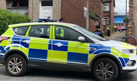 A police car in the foreground with officers stood in an alleyway behind a cordon