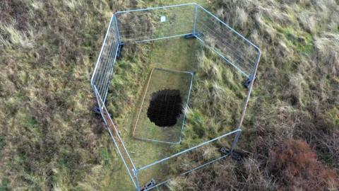 A hole in the ground, from above, surrounded by six metal fences. The hole is in the middle of a grassy area which is surrounded by longer rough grass like the type found in sand dunes. A fence panel is resting on top of the hole that is in the middle of the image.
