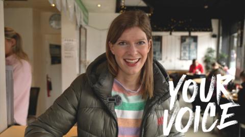 Colletta Smith smiles to the camera from a cafe. The picture has the words "your voice" written on the right.