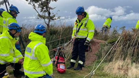 Seven members of Mullacott Coastguard Search and Rescue are pictured. They are positioned on the cliff top with one member wearing a harness and it looks as though he is preparing to go down the cliff face. All the members are in high-viz jackets and black trousers.