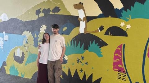 Vinny Lynch and Gemma Pasmore are standing in front of the mural smiling at the camera. He has his arm around her shoulders. He has black hair and a black beard and she has shoulder length brown hair. They are both wearing white T-shirts and she is wearing a skirt and he is wearing cargo trousers.  The mural shows a stoat standing up on its back legs which is two thirds the height of the students. There is a swan on the lake and on the shore a nest with three large eggs in it.