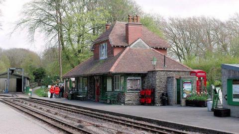 A picture taken from the outside of Woody Bay Station. There is a small railway line with a building next to it. There is people stood on the platform in the distance.