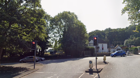 Three traffic lights on a road with trees in the background. There is a blue car visible on the junction