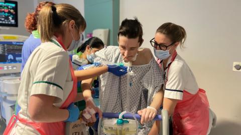 A patient in the intensive care unit is helped by nurses and occupational therapists wearing masks and aprons 