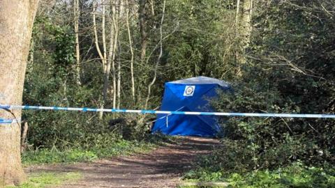 A police cordon attached to a tree in the middle of a wooded area. A blue police tent can be seen further down the footpath next to some trees.