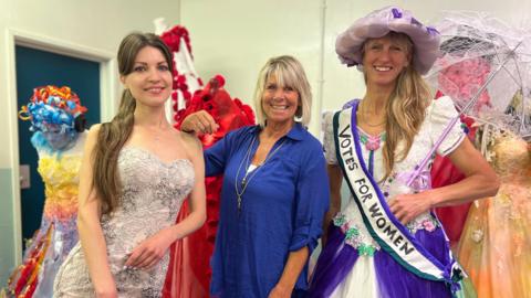 Three project participants showing off some of the dresses. The woman on the right is wearing a white and purple dress with a 'votes for women' sash and carrying a parasol.