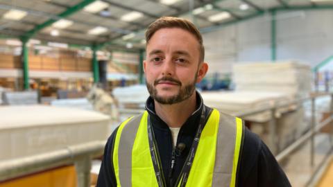 A 33 year old man with a beard and moustache half-smiles at the camera whilst wearing a high-vis tabard. He stands on a low gantry in a warehouse with piles of cut wood behind him.
