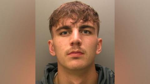 A mugshot of Kian Rowe. He has short brown hair, shaved on the sides and longer on the top, with some strands of hair coming down on to his forehead. He has blue eyes and a sparse moustache. He is wearing a dark grey hoodie and is looking serious.