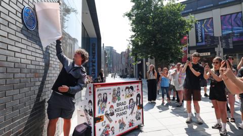 Brad Kella is standing, and pulls a white cover from a blue plaque as people watch and take photographs on their phones beside a decorated piano. It is sunny day. 