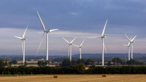 Seven large white wind turbines tower over a sunny countryside scene, including a golden harvested wheat or corn field in the foreground.