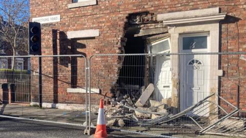 An end terrace house which has its front door caved in. There is a large hole to the left of the white plastic door with a pile of rubble beside it. The area has been blocked off with metal fences and there is a white and orange traffic cone in front of the fence. To the left of the door is a bricked up window and there is a traffic light in front of it which is off.
