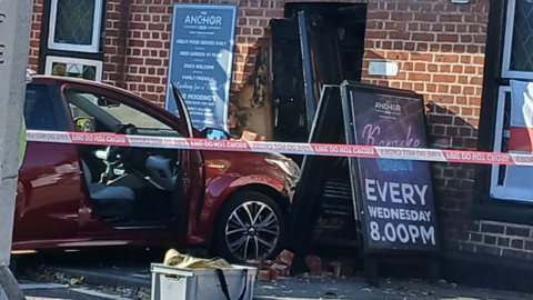 A crimson coloured vehicle can be seen after it has crashed into a pub. Some of the brickwork has been damaged and has fallen away.