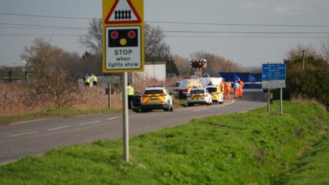 There are lots of emergency vehicles on a road near a level crossing. There is a blue tent next to the crossing and trees.