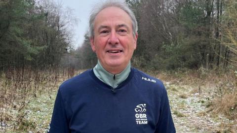 Mark Burtonshaw faces the camera, dressed in athletic gear. He wears a light green thermal long-sleeved top, and navy blue t-shirt with a white GBR logo on the chest. He has short grey-white hair and stands against the backdrop of a frosty woodland.