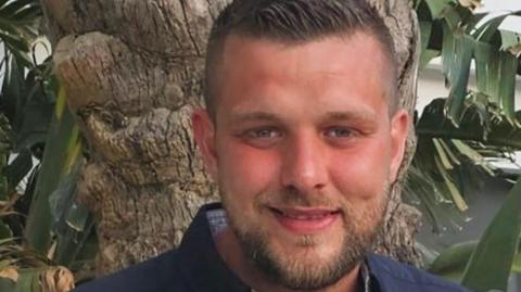 Close-up of a man with short brown hair and beard standing in front of a palm tree, smiling.