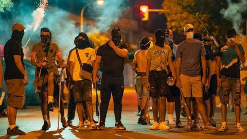 A group of around a dozen men, mostly wearing shorts, t-shirts and trainers, wear masks and balaclavas as they stand around on a street at night time. Some hold baseball bats, sticks and chains, while a flare is lit in the background
