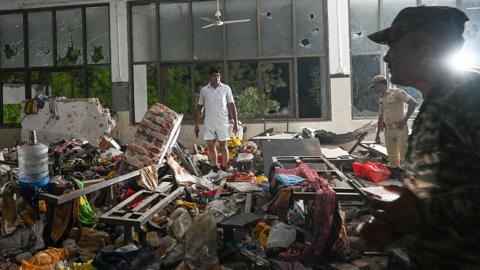People stand amid a chaotic scene of debris within the doctors' mess hall after the crash. It is clearly dark outside, meaning it is several hours since the incident