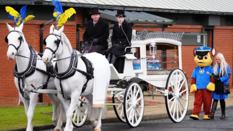 Two bright white horses with blue and yellow feathers pull a white carriage, carrying the small blue coffin of Hope Gordon. Behind the carriage is a person dressed in a Paw Patrol costume