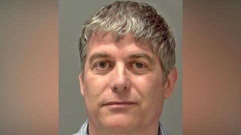 A police custody image of a man who is looking directly at the camera and slightly smiling. He has grey hair that slightly hangs over his forehead and he is wearing a blue shirt with white stripes on it. 