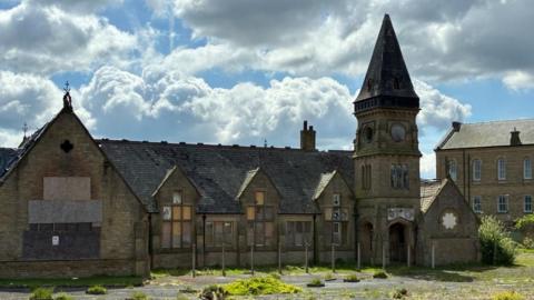 A Grade II listed school building made of light coloured stone, in a derelict state. The building is one-storey with a clocktower. Large windows are boarded up, and a extensive playground area is covered in grass and weeds.