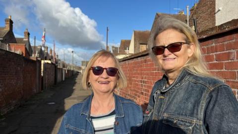 Jenny Butcher, standing on the left, and Linda Osborne. Both are wearing denim jackets, and sun glasses, and they both have blonde hair. They are standing in an alley - which stretches into the distance behind them - between two rows of terraced houses. 