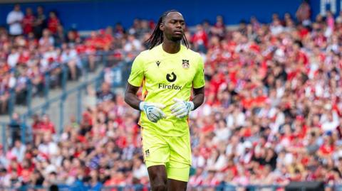 Arthur Okonkow jogging on the pitch wearing fluorescent yellow Wrexham kit and white goalkeeper gloves