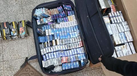 A case packed full of packs of cigarettes is held open by a hand covered with a black glove. The case is surrounded by other packs of cigarettes on the floor and a cardboard box of more cigarettes.