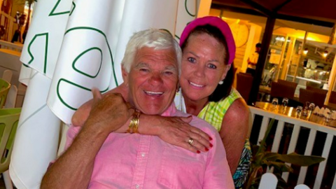 An elderly man wears a pink shirt and is sitting on a chair while a woman with a pink hairband and dark hair embraces him, both are smiling at the camera. 