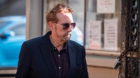 The image depicts a Chris Brain walking outdoors during the day. He is wearing a dark blazer and sunglasses, and has short hair along with facial hair. In the background, there is a blue car parked near a building that has several notices or signs posted on its window.