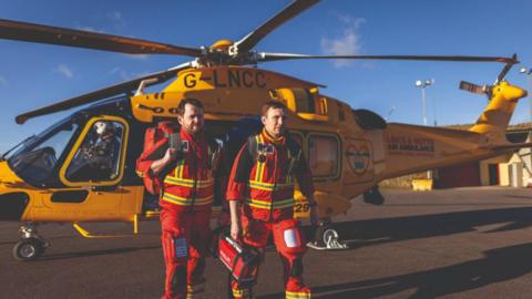 Two men dressed in red overalls with yellow fluorescent stripes on them are carrying paramedic items and walking away from a yellow Lincs & Notts Air Ambulance helicopter that is parked on tarmac.