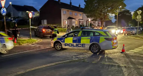 Two police cars on a road. There is police tape on sign posts. There is a car driving outside of the cordon and a person in police uniform directing them. There are houses in the background and cars with their headlights on. 