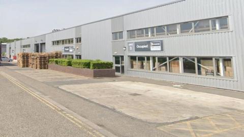 A view of three two-storey metal-clad industrial units with a road in front of them. There is a blue sky above.