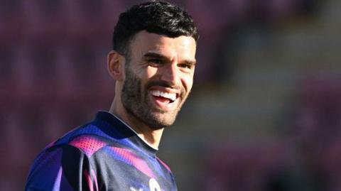 Conor Thomas smiles during a warm-up for Crewe Alexandra. He is wearing a multi-coloured top of black, purple and white.