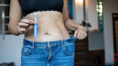 A woman celebrates her weight-loss progress by holding a injector in one hand and pulling on the waistband of jeans that no longer fit, showing her transformation.