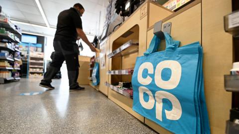 A blue bag-for-life with a big white 'Co-op' logo is seen hanging on a peg next to the checkout in a Co-op store with a worker in the background stacking shelves, in London in 2021.