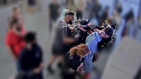 A video grab image issued by the Crown Prosecution Service 
from CCTV footage at Manchester Airport which has been played to jurors at Liverpool Crown Court. Three officers are standing around Mohammed Fahir Amaaz while his brother is standing behind them. One of the officers as his hand on the back of Amaaz's neck. 