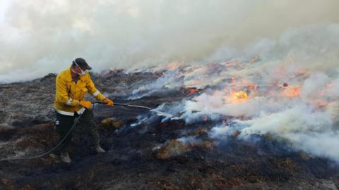 A person wearing a cap, face covering and a protective jacket sprays water on to flames burning across an area of moorland