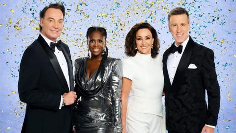 From left to right Craig Revel Horwood, Motsi Mabuse, Head Judge Shirley Ballas and Anton Du Beke can be seen standing in front of a blue background with glitter in the air around them. 
