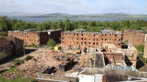 An aerial shot of a derelict red sandstone estate surrounded by trees with a river in the background - a number of the buildings are partly or fully demolished.