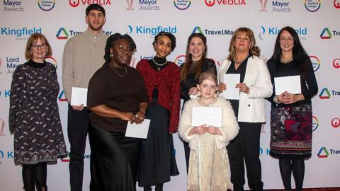 A group of eight people standing together on a red carpet in front of a backdrop covered with logos for the Lord Mayor Awards, Veolia, Kingfield, and TravelMaster. The group is dressed in smart and semi-formal clothing, with several individuals holding white envelopes or papers. Heidi stands at the front holding a sheet of paper.
