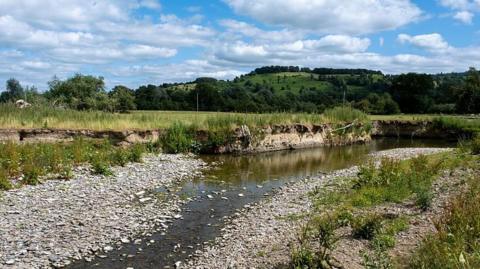 A mostly dried out river bed with some water in it in a landscape of green grass and hills in the background with a blue sky with white clouds above 